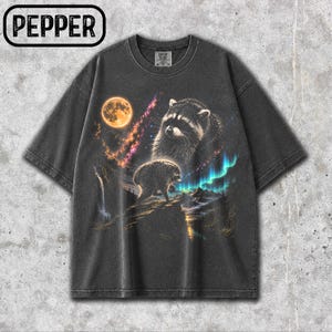 Puede incluir: Una camiseta gris carb&oacute;n de manga corta con un estampado gr&aacute;fico de un mapache sobre un acantilado rocoso bajo una luna llena y una nebulosa colorida. El dise&ntilde;o incluye una cascada, picos de monta&ntilde;as y la palabra PEPPER en un recuadro.