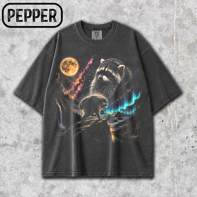 Puede incluir: Una camiseta gris carb&oacute;n de manga corta con un estampado gr&aacute;fico de un mapache sobre un acantilado rocoso bajo una luna llena y una nebulosa colorida. El dise&ntilde;o incluye una cascada, picos de monta&ntilde;as y la palabra PEPPER en un recuadro.