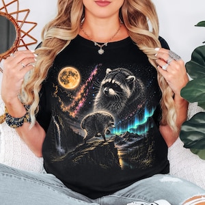 Puede incluir: Una camiseta negra con un estampado gr&aacute;fico de dos mapaches en un paisaje nocturno. El dise&ntilde;o incluye una luna llena grande, una nebulosa vibrante y luces de aurora boreal verde sobre una cadena monta&ntilde;osa con una cascada y un r&iacute;o.