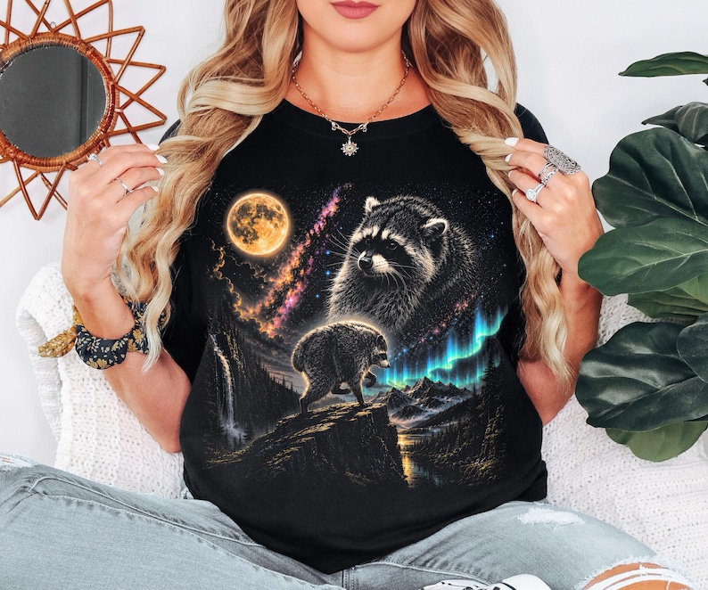 Puede incluir: Una camiseta negra con un estampado gr&aacute;fico de dos mapaches en un paisaje nocturno. El dise&ntilde;o incluye una luna llena grande, una nebulosa vibrante y luces de aurora boreal verde sobre una cadena monta&ntilde;osa con una cascada y un r&iacute;o.