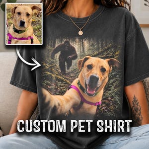 May include: A charcoal grey t-shirt featuring a graphic print of a tan dog wearing a pink harness and a black collar, appearing to take a selfie in a forest with a Bigfoot figure in the background. The text CUSTOM PET SHIRT is printed below.