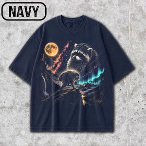 Puede incluir: Una camiseta azul marino de manga corta con un estampado gr&aacute;fico de dos mapaches en un paisaje nocturno. El dise&ntilde;o incluye una luna llena, una nebulosa, una cascada, monta&ntilde;as y auroras boreales. La palabra NAVY est&aacute; impresa en la esquina superior.