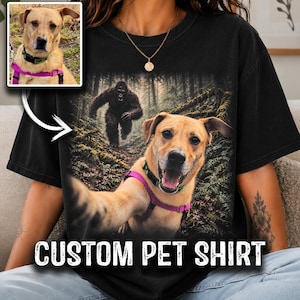 May include: A black t-shirt featuring a graphic print of a tan dog wearing a purple harness and a green collar, taking a selfie in a forest with a large dark creature in the background. A small inset photo shows the same dog in the same harness.