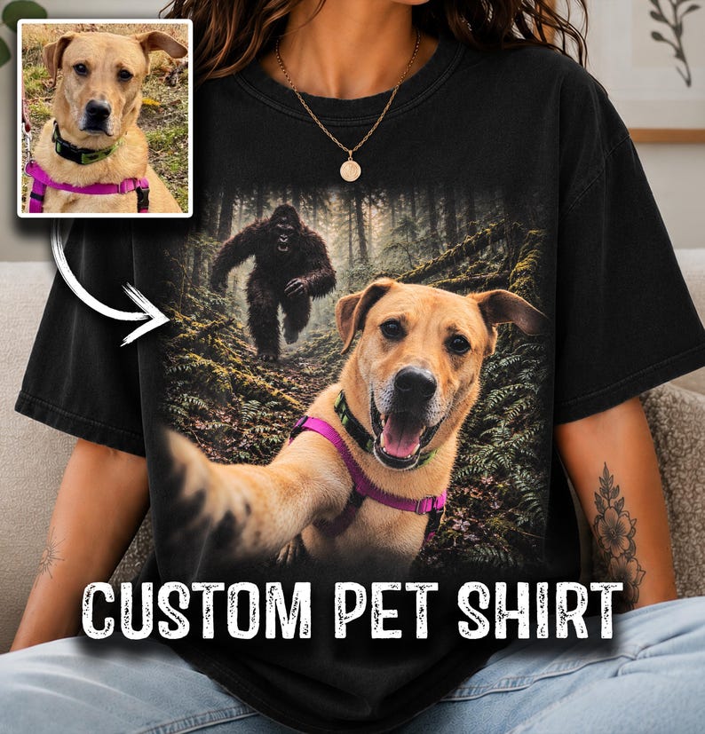 May include: A black t-shirt featuring a graphic print of a tan dog wearing a purple harness and a green collar, taking a selfie in a forest with a large dark creature in the background. A small inset photo shows the same dog in the same harness.