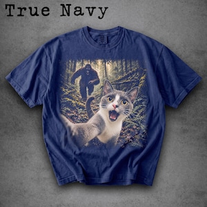May include: A True Navy short-sleeve t-shirt featuring a graphic print of a grey and white cat taking a selfie in a forest, with a large, dark, hairy creature resembling Bigfoot walking in the background among the trees and ferns.