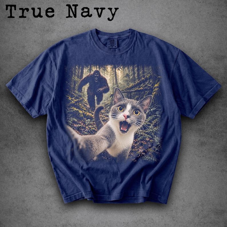 May include: A True Navy short-sleeve t-shirt featuring a graphic print of a grey and white cat taking a selfie in a forest, with a large, dark, hairy creature resembling Bigfoot walking in the background among the trees and ferns.