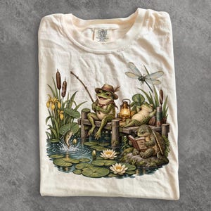 May include: A cream-coloured t-shirt featuring a graphic of a frog wearing a hat and fishing from a wooden dock, a frog resting near a lantern, and a turtle reading a book on a rock. A dragonfly flies above, and a fish jumps from the water.