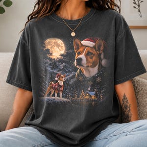 May include: A charcoal gray short-sleeve t-shirt featuring a graphic print of a Basenji dog wearing a Santa hat and a patterned sweater. The scene includes a full moon, a snowy mountain landscape, a small cabin, and a second smaller dog.
