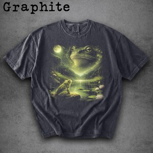 May include: Graphite-colored t-shirt featuring a detailed illustration of a frog in a mystical pond setting. The design includes a large frog, a smaller frog, a full moon, and water lilies. The shirt has a classic crew neck.