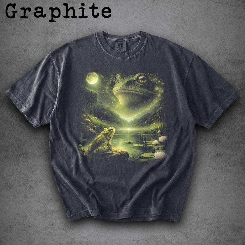 May include: Graphite-colored t-shirt featuring a detailed illustration of a frog in a mystical pond setting. The design includes a large frog, a smaller frog, a full moon, and water lilies. The shirt has a classic crew neck.