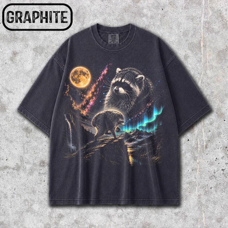 Puede incluir: Una camiseta gris carb&oacute;n con el texto GRAPHITE en un recuadro negro. El gr&aacute;fico muestra un mapache sobre un acantilado bajo una luna llena, con una nebulosa, una cascada y brillantes auroras boreales de color verde en el cielo.