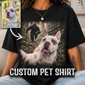 May include: A black t-shirt featuring a graphic print of a white and brown dog taking a selfie in a forest with a large dark creature resembling Bigfoot in the background. The text CUSTOM PET SHIRT is printed in white at the bottom of the shirt.