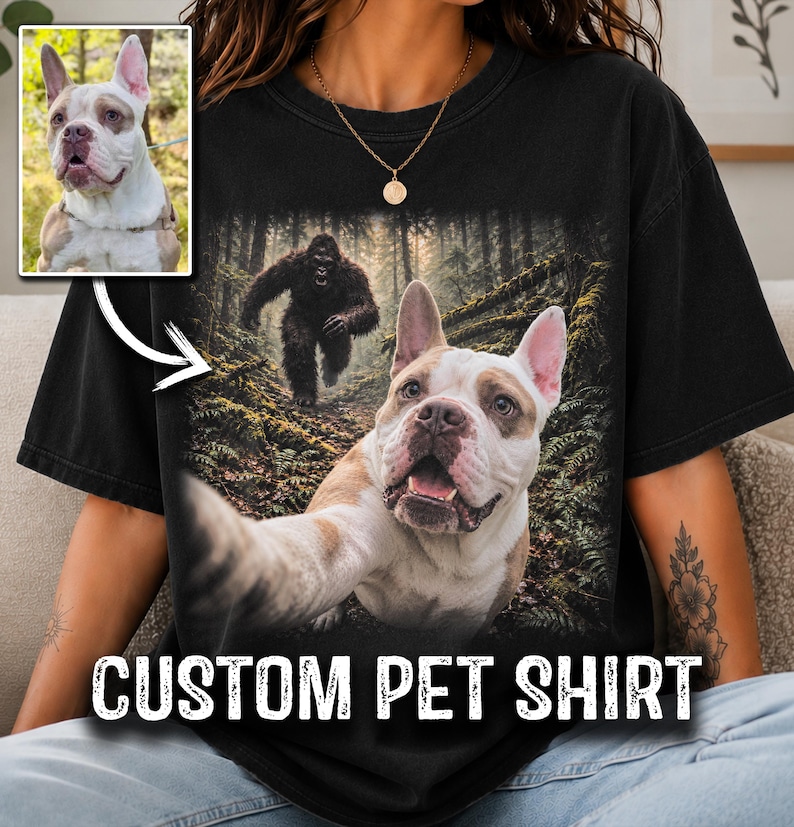 May include: A black t-shirt featuring a graphic print of a white and brown dog taking a selfie in a forest with a large dark creature resembling Bigfoot in the background. The text CUSTOM PET SHIRT is printed in white at the bottom of the shirt.
