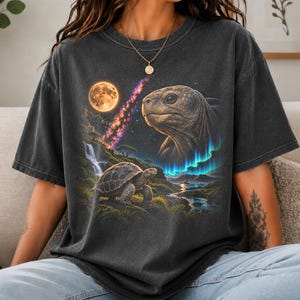 May include: A charcoal gray short-sleeve t-shirt featuring a graphic print of a large tortoise head in the sky above a landscape with a smaller tortoise, a waterfall, a glowing full moon, a colorful nebula, and vibrant blue aurora borealis lights.