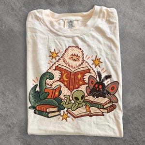 May include: A cream-colored t-shirt featuring a graphic illustration of a white yeti, a green dinosaur, a green alien, and a moth-like creature all reading books. The design includes orange stars and sun motifs, with the Comfort Colors brand label visible at the neck.