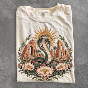 May include: A cream-coloured t-shirt featuring a graphic print of a cobra snake centred in front of ancient stone ruins and a sun. The design is framed by lotus flowers and green leaves. The shirt has a Comfort Colors brand label at the neck.