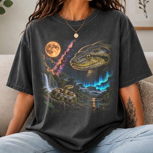 May include: A charcoal grey short-sleeved t-shirt featuring a graphic print of a large snake head and a coiled snake in a jungle landscape. The scene includes a full moon, a waterfall, and vibrant blue aurora borealis lights against a dark sky.