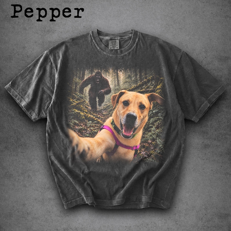 May include: A charcoal grey t-shirt featuring a graphic print of a light brown dog wearing a pink harness taking a selfie in a forest. In the background, a dark, hairy creature resembling a Bigfoot is visible among the trees. The word Pepper is printed in black text at the top left corner of the image.