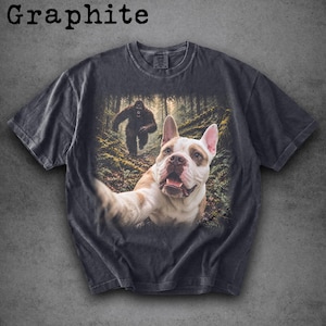 May include: A graphite-coloured short-sleeve t-shirt featuring a graphic print of a French Bulldog taking a selfie in a forest with a Bigfoot figure in the background. The shirt has a crew neckline and a faded, vintage-inspired wash finish.