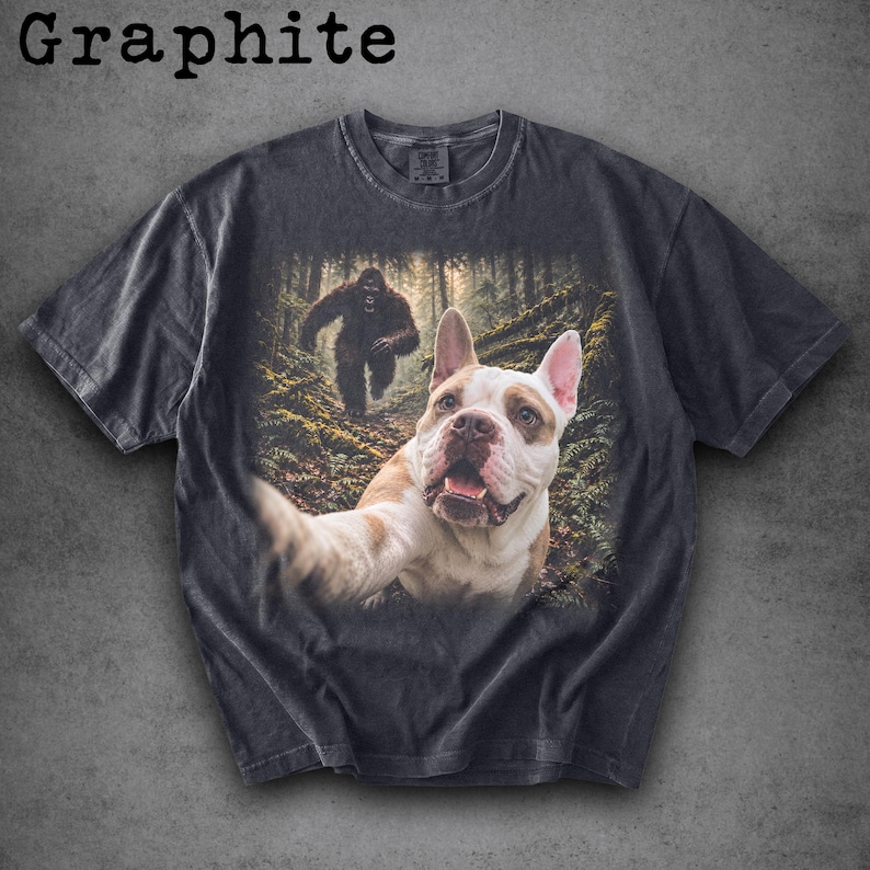 May include: A graphite-coloured short-sleeve t-shirt featuring a graphic print of a French Bulldog taking a selfie in a forest with a Bigfoot figure in the background. The shirt has a crew neckline and a faded, vintage-inspired wash finish.