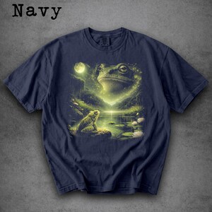 May include: Navy blue t-shirt featuring a detailed illustration of a frog scene. The design includes a large frog head, a smaller frog, a moon, and a pond with lily pads. The word "Navy" is printed at the top.