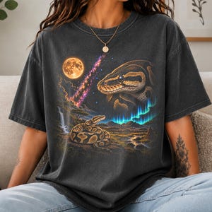May include: A charcoal grey short-sleeve t-shirt featuring a graphic print of a large snake head in the upper right, a smaller coiled snake on a rocky landscape below, a full moon, a purple nebula, and bright blue aurora borealis lights.