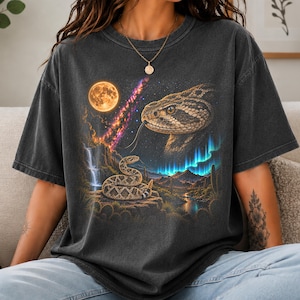 May include: A charcoal grey short-sleeve t-shirt featuring a graphic print of a desert landscape with a coiled rattlesnake, a large moon, a waterfall, cacti, and a giant snake head in the sky with a colourful nebula and aurora borealis.