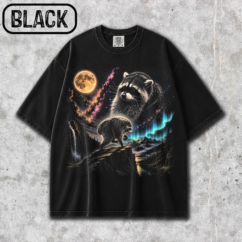 Puede incluir: Una camiseta negra de manga corta con un estampado gr&aacute;fico de un mapache sobre un acantilado rocoso bajo una luna llena y una nebulosa. El dise&ntilde;o incluye una cascada, un paisaje monta&ntilde;oso y luces de aurora boreal en tonos azules y verdes.
