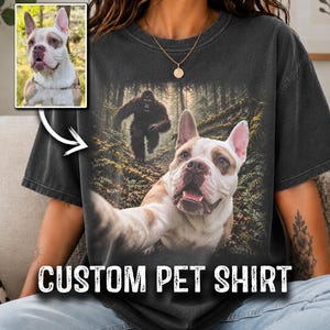 May include: A charcoal grey short-sleeve t-shirt featuring a graphic print of a white and brown dog taking a selfie in a forest with a large dark creature in the background. The shirt includes the text CUSTOM PET SHIRT at the bottom.