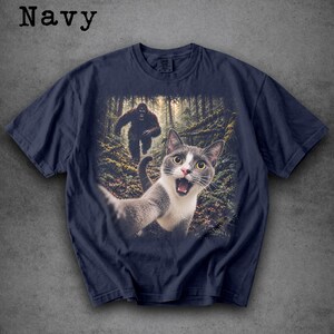 May include: A navy blue short-sleeve t-shirt featuring a graphic print of a grey and white cat taking a selfie in a forest with a large, dark, hairy creature resembling Bigfoot walking in the background among the trees and ferns.