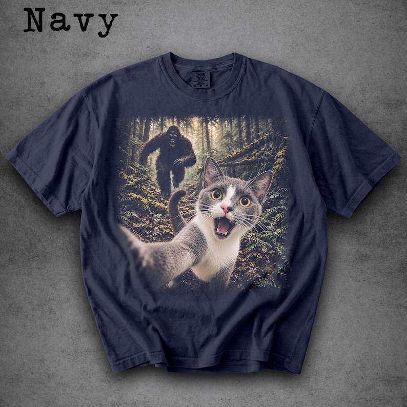 May include: A navy blue short-sleeve t-shirt featuring a graphic print of a grey and white cat taking a selfie in a forest with a large, dark, hairy creature resembling Bigfoot walking in the background among the trees and ferns.