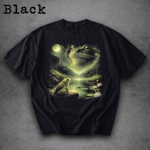 May include: Black t-shirt featuring a detailed illustration of a frog scene. The design includes a large frog face, a smaller frog, a moon, and a pond with lily pads. The word "Black" is printed at the top.