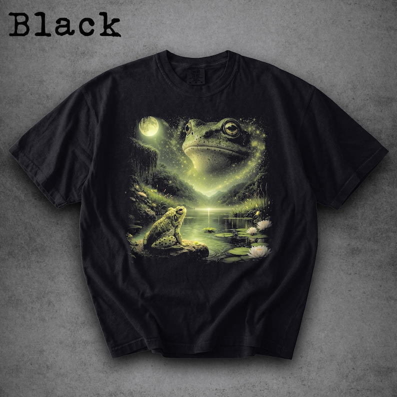 May include: Black t-shirt featuring a detailed illustration of a frog scene. The design includes a large frog face, a smaller frog, a moon, and a pond with lily pads. The word "Black" is printed at the top.