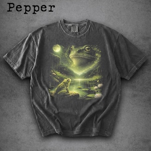 May include: Dark grey t-shirt featuring a detailed illustration of a frog in a serene pond setting under a full moon. The design includes a large frog face and a smaller frog on a rock, with the word "Pepper" at the top.