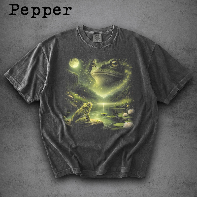 May include: Dark grey t-shirt featuring a detailed illustration of a frog in a serene pond setting under a full moon. The design includes a large frog face and a smaller frog on a rock, with the word "Pepper" at the top.