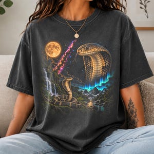 May include: A charcoal grey short-sleeve t-shirt featuring a graphic print of two cobras in a jungle landscape with a waterfall, a full moon, a nebula, and aurora borealis. The design includes a large cobra head and a smaller coiled cobra.
