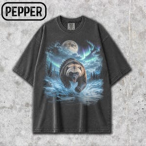 May include: A charcoal grey t-shirt featuring a graphic print of a brown bear running through water. The background shows a full moon, a forest of pine trees, and vibrant blue and purple aurora borealis lights. The word PEPPER is in the top corner.