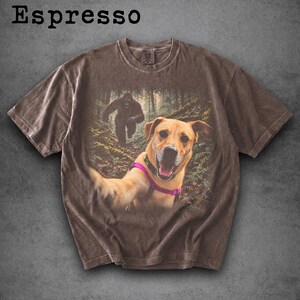May include: An espresso-coloured short-sleeve t-shirt featuring a graphic print of a golden dog wearing a pink harness taking a selfie in a forest, with a large dark creature resembling Bigfoot walking in the background behind the dog.