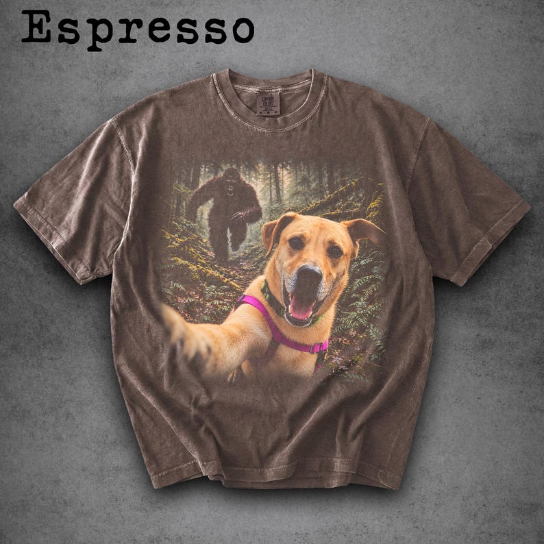 May include: An espresso-coloured short-sleeve t-shirt featuring a graphic print of a golden dog wearing a pink harness taking a selfie in a forest, with a large dark creature resembling Bigfoot walking in the background behind the dog.