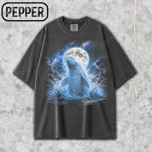 Puede incluir: Una camiseta de manga corta gris carbón con un estampado gráfico de una beluga blanca emergiendo de agua azul frente a una gran luna llena y un cielo estrellado. La palabra PEPPER está impresa en un recuadro negro en la parte superior izquierda.