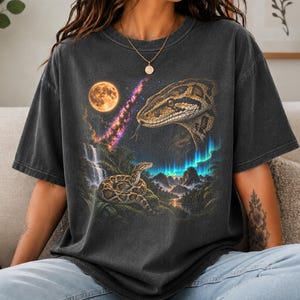 May include: A charcoal grey short-sleeve t-shirt featuring a graphic print of a large snake head in the upper right and a smaller coiled snake on a rock in a landscape with a waterfall, a full moon, a nebula, and aurora borealis lights.