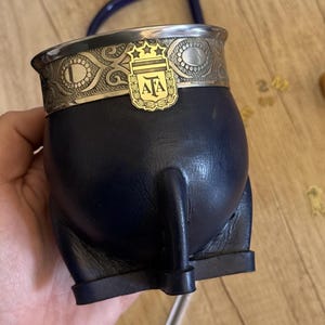 Puede incluir: Un mate forrado en cuero azul oscuro con un borde de metal plateado que presenta un patrón en relieve. Un emblema dorado con las letras AFA y tres estrellas está fijado en la parte frontal. La base cuenta con cuatro pequeñas patas de cuero.