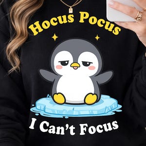 May include: Black sweatshirt with a graphic of a penguin on an ice floe. The text "Hocus Pocus" is above the penguin, and "I Can't Focus" is below. The penguin is gray, white, and yellow.