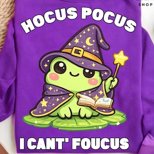 May include: Purple sweatshirt with the text "Hocus Pocus" above a cartoon frog wearing a witch hat and cape. The frog is holding a wand and a book. The text "I Can't Foucus" is below the frog.