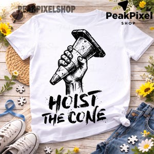 Pu&ograve; includere: T-shirt bianca con grafica nera di una mano che tiene un cono stradale. Il testo "HOIST THE CONE" &egrave; sotto la grafica. La maglietta &egrave; esposta con jeans blu, scarpe da ginnastica bianche e girasoli.