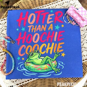 May include: A blue t-shirt featuring the text HOTTER THAN A HOOCHIE COOCHIE in pink, orange, and yellow. Below the text is a green frog wearing sunglasses, lounging on a colorful inflatable pool float in blue water, accented by small stars.