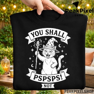 May include: Black sweatshirt featuring a white illustration of a cat wearing a wizard hat and holding a staff. The text "YOU SHALL" is above the cat, and "PSPSPS! NOT" is below. The design includes stars and crescent moons.