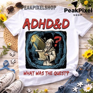 Puede incluir: Una camiseta blanca con el texto rojo ADHD&D en la parte superior y el texto rojo WHAT WAS THE QUEST? en la parte inferior. El gráfico central muestra a un mago barbudo en una cueva sosteniendo un pergamino, iluminado por un rayo de luz, con un signo de interrogación rojo.