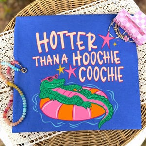 May include: A royal blue t-shirt featuring a graphic of a green alligator wearing pink sunglasses, lounging on a pink and orange striped pool float. The text reads "HOTTER THAN A HOOCHIE COOCHIE" in light yellow letters, accented by pink and yellow stars.