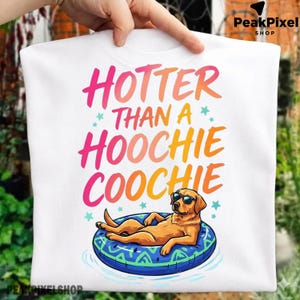 May include: A white t-shirt featuring a graphic of a golden dog wearing sunglasses while lounging on a blue patterned pool float. The text above the dog reads HOTTER THAN A HOOCHIE COOCHIE in a vibrant pink, orange, and yellow gradient font.